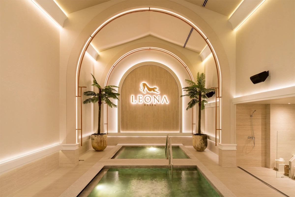 A symmetrical, luxury men’s onsen featuring twin mineral pools under a grand archway. The minimalist, cream-toned space is anchored by the "LEONA ONSEN" jaguar logo and framed by ambient lighting and palm trees, creating a sophisticated sanctuary for gentlemen.